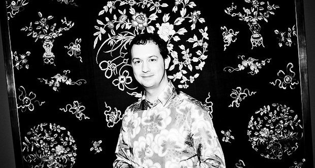 Retrato en blanco y negro de un hombre sonriendo, vestido con una camisa de flores, frente a un fondo decorativo.