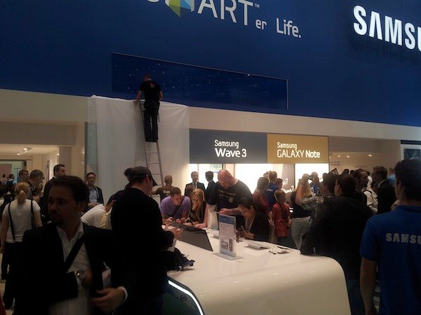 Ein belebter Messestand von Samsung mit Leuten, die sich um Geräte wie das Galaxy Note und Wave 3 versammeln.